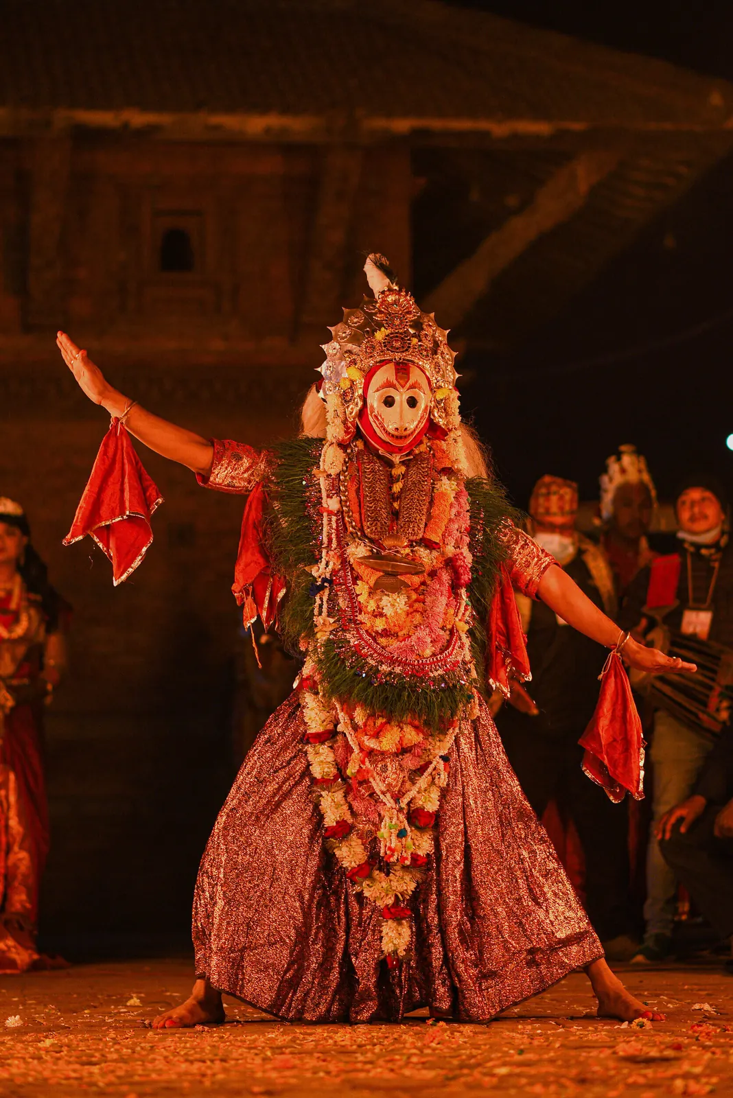 A dancer in a red monkey mask and flower crown spreads his arms