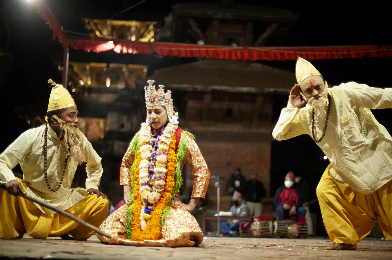 Two actors as sages flank a crowned dancer seated on the stage