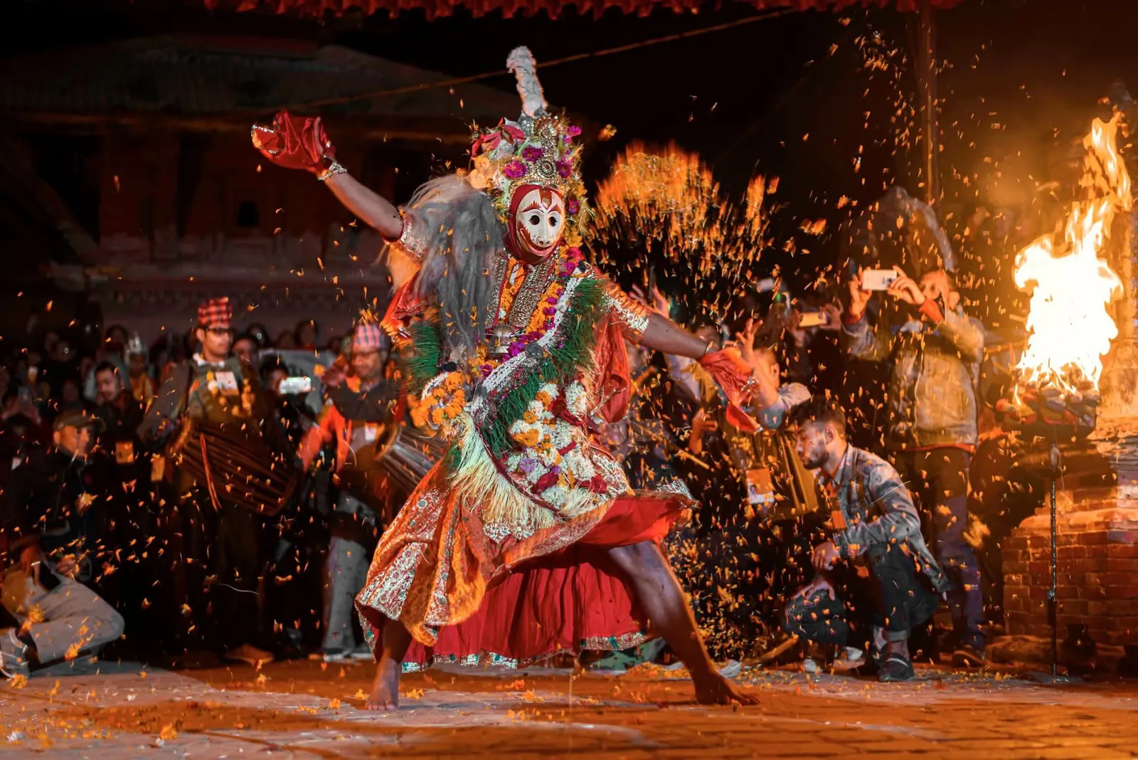A masked dancer strikes a pose as marigold petals fly beside a fire torch